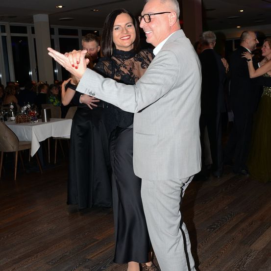 Ein älterer Mann und eine Frau tanzen lächelnd zusammen auf einem Holzboden. Sie sind von anderen Menschen in formeller Kleidung umgeben. Im Hintergrund befindet sich ein Tisch mit weißer Tischdecke und Stühlen.