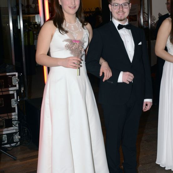 Eine Frau in einem weißen Kleid und ein Mann in einem schwarzen Anzug und einer Fliege stehen zusammen. Die Frau hält einen kleinen Blumenstrauß. Sie befinden sich bei einer formellen Veranstaltung.