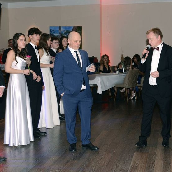 Eine formelle Veranstaltung, bei der sich Menschen versammelt haben. Ein Mann in formeller Kleidung spricht in ein Mikrofon, während ein anderer Mann vor ihm steht und etwas von einem Papier liest.