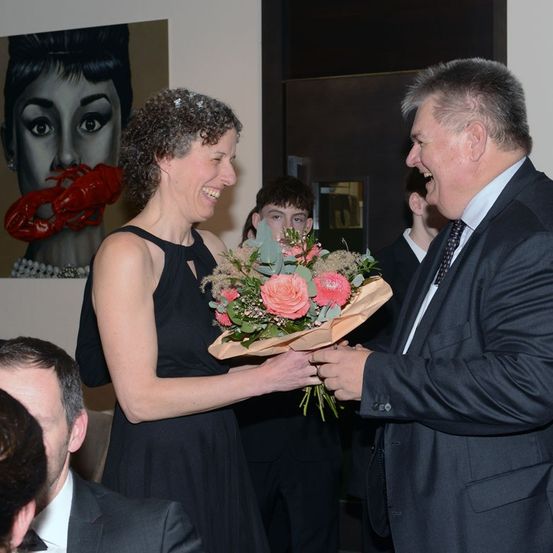 Ein Mann und eine Frau tauschen Blumen bei einer Veranstaltung im Innenbereich aus. Die Frau lacht, während der Mann lächelt. Hinter ihnen ist ein Bild einer Frau mit einem Hummer zu sehen.