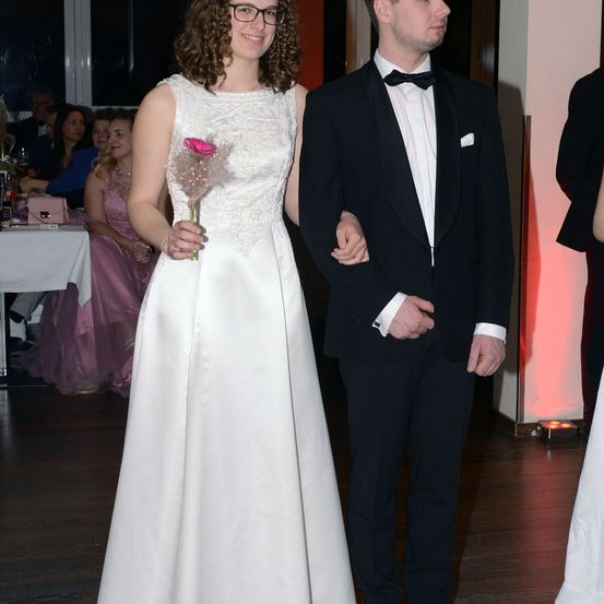 Ein Paar steht bei einer Veranstaltung, lächelt und die Frau hält eine rosa Blume. Hinter ihnen sitzen andere an Tischen und der Raum ist sanft beleuchtet.