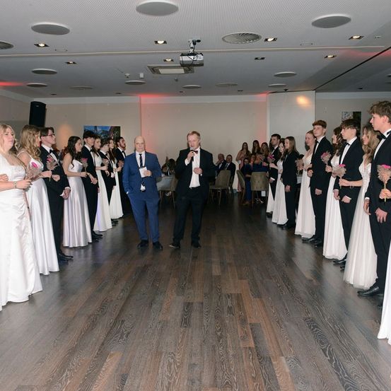 Eine Gruppe von Menschen steht in einem Raum, einige halten Blumen. Zwei Männer stehen vorne, einer mit einem Mikrofon, wahrscheinlich bei einer Rede. Der Raum hat Holzböden und eine Decke mit Lichtern.