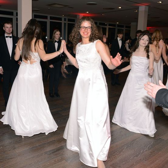 Eine Gruppe von Frauen in weißen Kleidern tanzt auf einem Holzboden, während einige Menschen zuschauen.