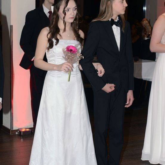 Eine junge Frau in einem weißen Kleid, die eine rosa Blume hält, steht mit einem Mann in einem schwarzen Frack bei einer formellen Veranstaltung. Sie schauen nach vorne. Hinter ihnen sind ein Mann und eine Frau in formeller Kleidung teilweise zu sehen. Der Hintergrund besteht aus einem roten Licht und einem Tisch mit weißer Tischdecke.