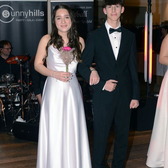 Ein junges Paar steht zusammen, das Mädchen hält eine rosa Blume, beide sind formell gekleidet. Hinter ihnen wirbt ein Banner für 'Sunnyhills Party.' Ein Musiker spielt Schlagzeug im Hintergrund.