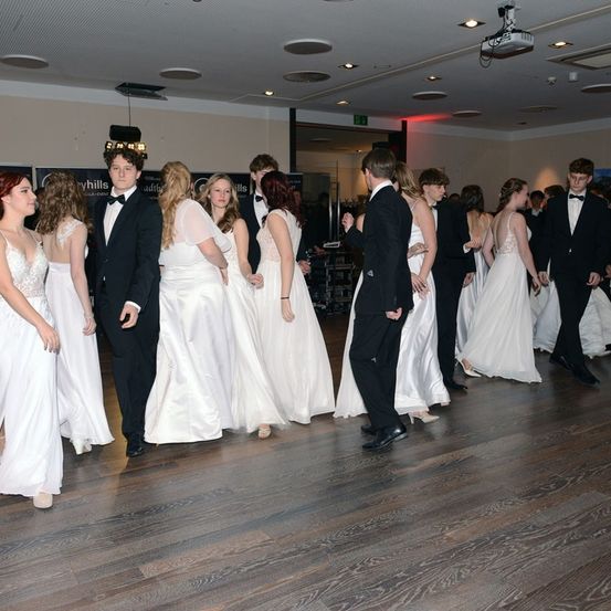 Eine Gruppe junger Menschen in formeller Kleidung, möglicherweise für einen Abschlussball, steht in einer Reihe auf einem Holzboden. Sie alle schauen nach vorne.