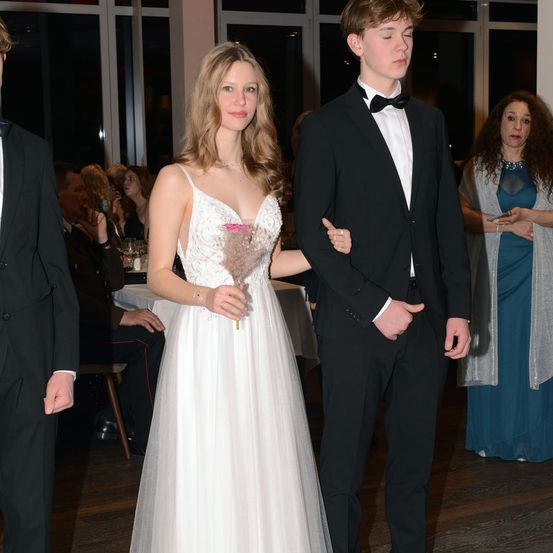 Ein junges Paar, vermutlich bei einer Hochzeit, geht zusammen, wobei das Mädchen eine rosa Blume hält. Dahinter beobachten ein Mann in Anzug und eine Frau in einem blauen Kleid.