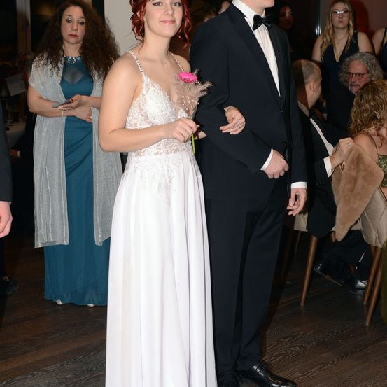 Eine Frau in einem weißen Kleid und ein Mann in einem schwarzen Anzug stehen bei einer Veranstaltung zusammen. Die Frau hält eine rosa Rose. Hinter ihnen sitzen und stehen Menschen an einem Tisch.