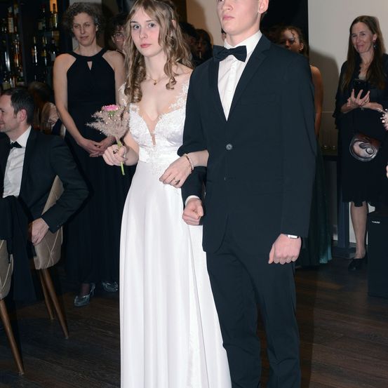 Ein junges Paar in formeller Kleidung, möglicherweise bei einer Hochzeitsfeier, wobei die Frau eine rosa Blume hält. Dahinter stehen und unterhalten sich mehrere Personen, vermutlich Gäste.