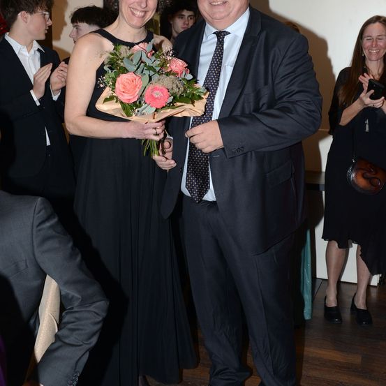 Eine Frau in einem schwarzen Kleid hält einen Blumenstrauß und steht neben einem Mann in einem Anzug. Sie befinden sich bei einer Veranstaltung, mit mehreren Personen hinter ihnen.