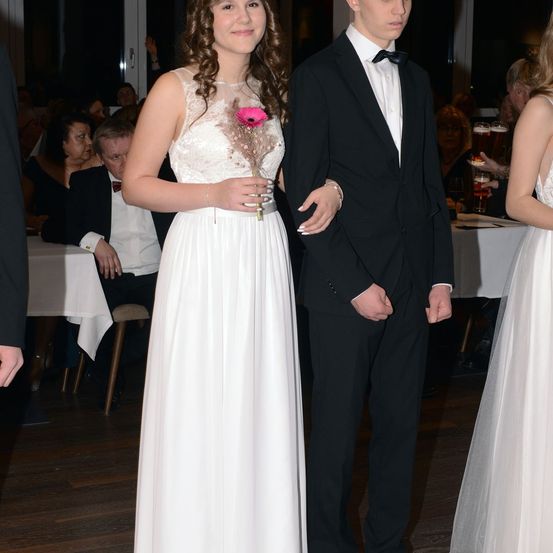 Eine junge Frau in einem weißen Kleid, die eine rosa Blume hält, steht neben einem Mann in einem schwarzen Anzug und Fliege. Dahinter sitzen Menschen an Tischen.