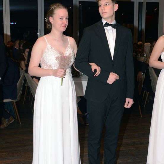 Ein junges Paar steht zusammen, die Frau in einem weißen Kleid und der Mann in einem schwarzen Anzug, lächelt für die Kamera mit anderen im Hintergrund.