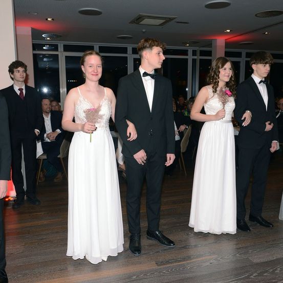 Eine Gruppe junger Menschen in formeller Kleidung, möglicherweise bei einem Abschlussball oder einer formellen Veranstaltung. Zwei Frauen halten Blumen, während ein Mann zwischen ihnen steht.