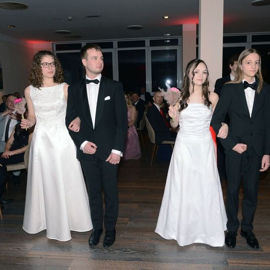 Vier Personen, zwei Männer und zwei Frauen, sind in formeller Kleidung gekleidet. Sie halten rosa Blumensträuße. Sie stehen auf einem Holzboden. Hinter ihnen sitzen mehrere Personen auf Stühlen.