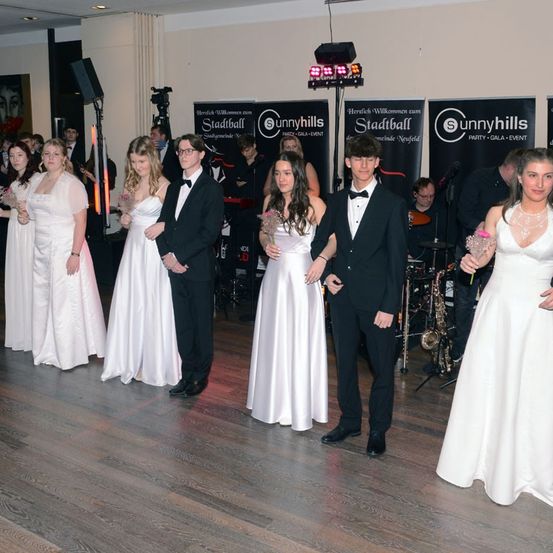 Mehrere Menschen in formeller Kleidung stehen auf einer Tanzfläche. Sie scheinen sich bei einer Schulveranstaltung zu befinden. Dahinter ist eine Bühne aufgebaut, mit Musikinstrumenten und Lautsprechern.