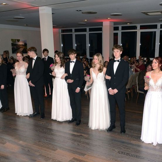 Eine Gruppe junger Menschen in formeller Kleidung steht auf einem Holzboden, möglicherweise bei einem Abschlussball oder einer formellen Veranstaltung. Sie lächeln und blicken nach vorne. Hinter ihnen stehen andere Leute, einige sitzen auf Stühlen. Der Raum hat eine Decke mit Lichtern und eine Glaswand.
