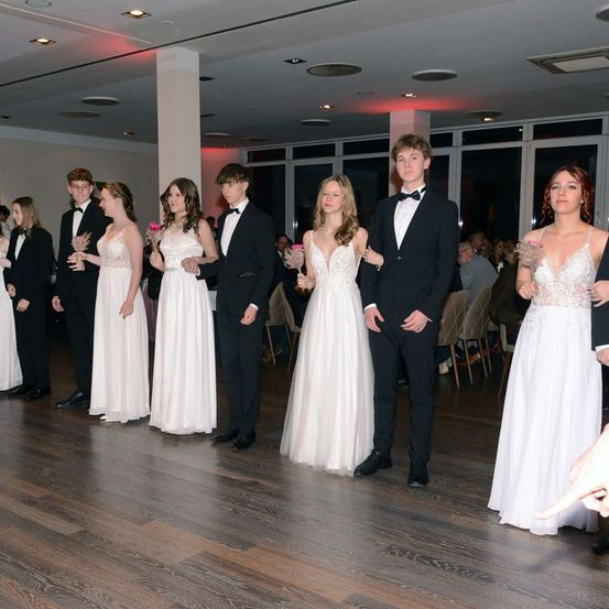 Eine Gruppe junger Menschen in formeller Kleidung steht auf einem Holzboden, möglicherweise bei einem Abschlussball oder einer ähnlichen Veranstaltung. Sie halten Blumen in den Händen.