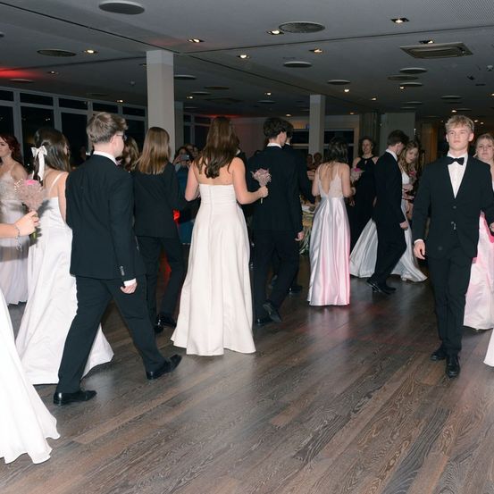 Mehrere Paare in formeller Kleidung tanzen auf einem hölzernen Tanzboden, einige halten Blumen. Der Raum ist mit roten und weißen Lichtern beleuchtet, und die Decke hat mehrere Lichter.