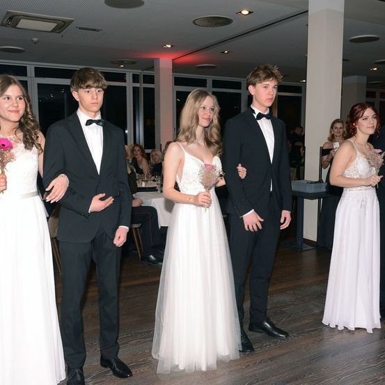 Fünf junge Erwachsene, drei Frauen und zwei Männer, in formeller Kleidung stehen zusammen auf einem Holzboden und halten Blumen. Hinter ihnen sitzen andere Gäste an Tischen.