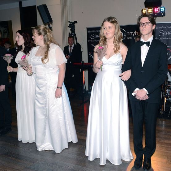 Drei Frauen in weißen Kleidern und ein Mann in einem schwarzen Anzug stehen auf einer Tanzfläche, wahrscheinlich bei einer Schulveranstaltung. Dahinter spielt ein Mann auf einem Keyboard und eine Kamera ist aufgestellt.