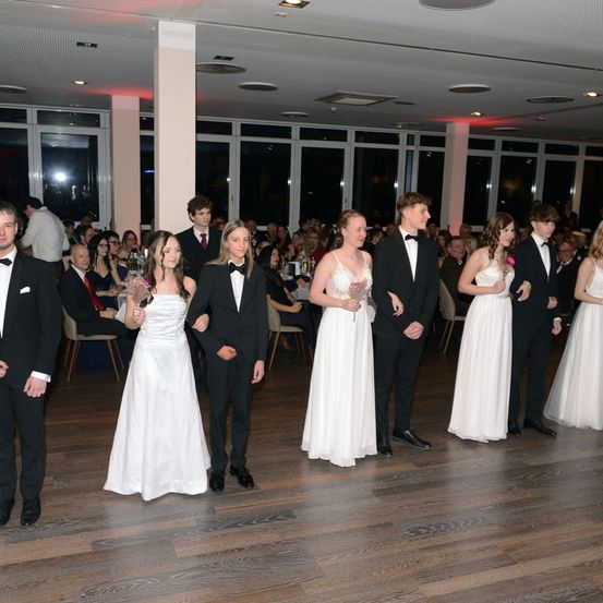 Eine Gruppe von Menschen, möglicherweise bei einer Hochzeit, steht in formeller Kleidung. Frauen in weißen Kleidern halten Blumen, während Männer Anzüge tragen. Die Umgebung umfasst einen Holzboden und Tische mit sitzenden Gästen.