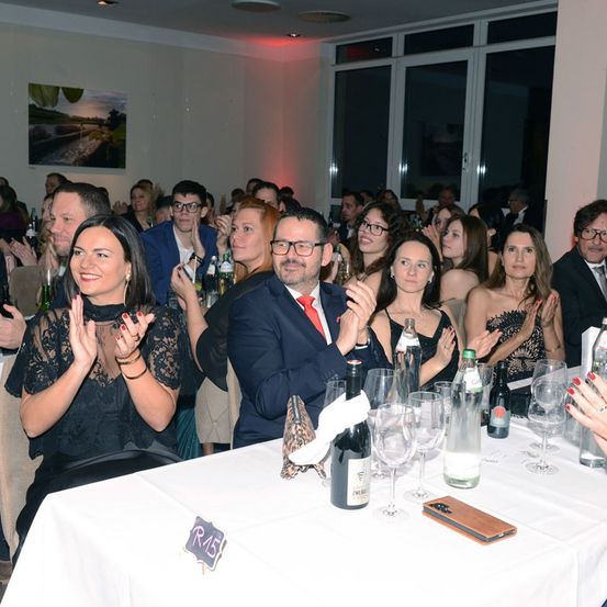 Eine große Gruppe von Menschen sitzt an einem Banketttisch in einem Bankettsaal. Der Tisch ist mit Weingläsern, Flaschen und Tellern gedeckt. Alle klatschen.