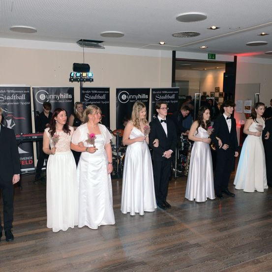 Eine Gruppe von Frauen in weißen Kleidern und Männern in Anzügen steht zusammen in einem Raum, möglicherweise bei einer Hochzeit. Im Hintergrund sind Musikinstrumente vorhanden.