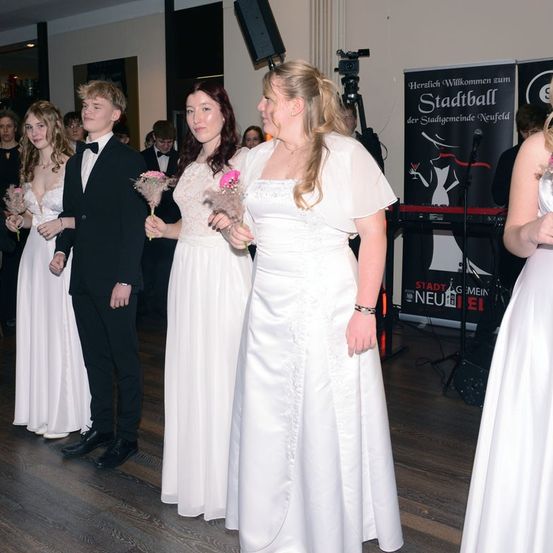 Eine Gruppe junger Menschen, möglicherweise auf einem Abschlussball, gekleidet in weißen Kleidern und schwarzen Anzügen, stehen auf einem Holzboden. Hinter ihnen befindet sich ein Banner mit Text und einem Bild einer Frau.