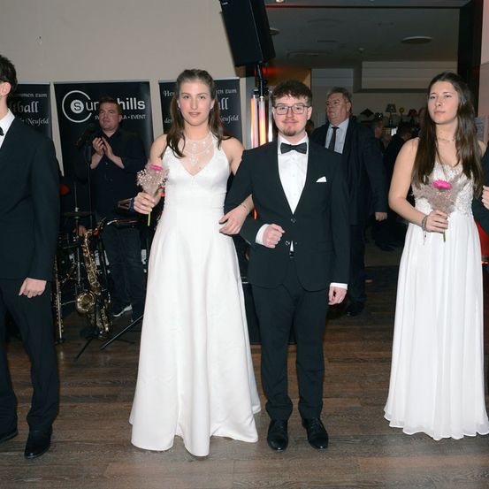 Drei junge Menschen in formeller Kleidung stehen bei einer Party zusammen. Ein Mann und eine Frau in weißen Kleidern und ein Mann im schwarzen Frack. Ein Saxophonist ist im Hintergrund zu sehen.