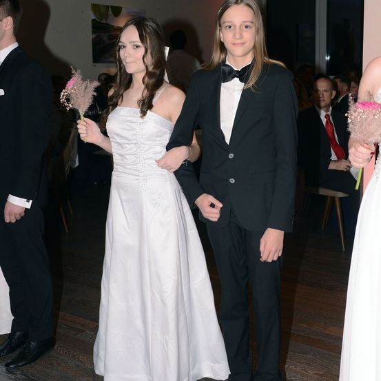 Ein Paar in formeller Kleidung steht in einem Raum. Die Frau hält eine rosa Blume. Hinter ihnen sitzen Leute auf Stühlen. Der Mann trägt einen schwarzen Anzug und eine Fliege.