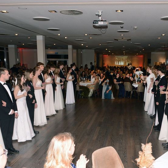 Eine große Gruppe von Menschen in formeller Kleidung versammelt sich in einem Raum. Einige halten Blumen. Eine Projektor ist an der Decke montiert. Einige Leute sitzen an einem Tisch. Eine Frau in einem blauen Kleid sitzt.