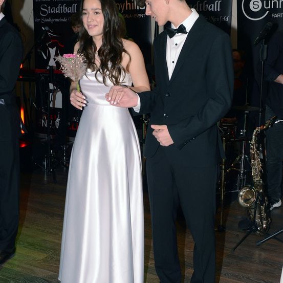 Ein Paar in formeller Kleidung steht lächelnd zusammen. Die Frau hält einen kleinen Blumenstrauß und der Mann hält ihre Hand. Hinter ihnen spielt eine Band mit Instrumenten auf einer Bühne.