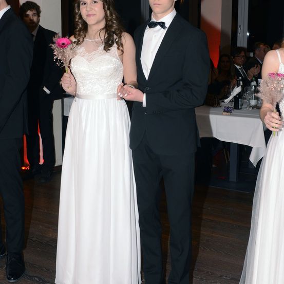 Ein junges Paar, wahrscheinlich auf einem Abschlussball, steht eng beieinander und lächelt. Das Mädchen hält eine rosa Blume und trägt ein weißes Kleid. Der Junge trägt einen schwarzen Frack mit einer Schleife. Sie stehen vor einem Tisch mit Flaschen und Servietten.