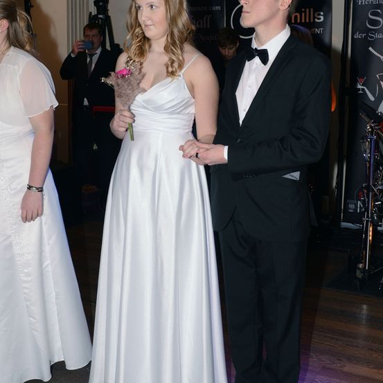 Ein Paar steht bei einer Veranstaltung zusammen und hält Händchen. Die Frau hält eine Blume. Hinter ihnen macht ein Mann ein Foto mit einer Kamera. Die Umgebung scheint eine formelle Veranstaltung zu sein.