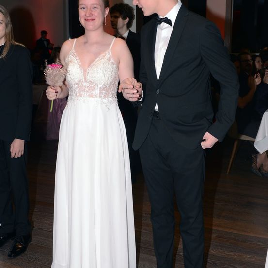 Ein Paar bei einer formellen Veranstaltung, die Frau in einem weißen Kleid, der Mann in einem schwarzen Anzug, halten Händchen, mit Leuten im Hintergrund.