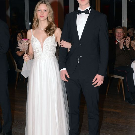 Ein junges Paar steht in formeller Kleidung bei einer Veranstaltung. Die Frau trägt ein weißes Kleid mit einer rosa Blume, und der Mann trägt einen schwarzen Anzug mit Fliege. Sie befinden sich in einem Raum mit Holzboden und sitzenden Gästen.