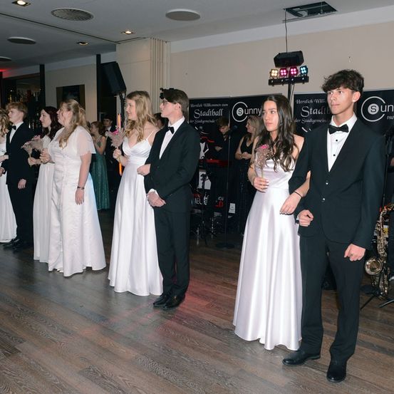 Eine Gruppe junger Erwachsener in formeller Kleidung steht zusammen, möglicherweise bei einem Abschlussball oder ähnlichem Event. Sie halten Blumen und stehen auf einem Holzboden.