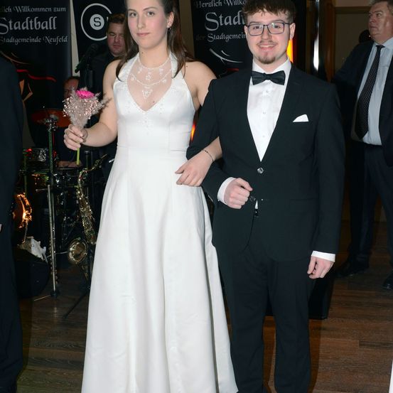 Ein junges Paar in formeller Kleidung bei einer Veranstaltung, wobei die Frau Blumen hält und der Mann eine Brille trägt. Dahinter spielt eine verschwommene Band auf der Bühne mit einem Schild, das Stadtball liest.