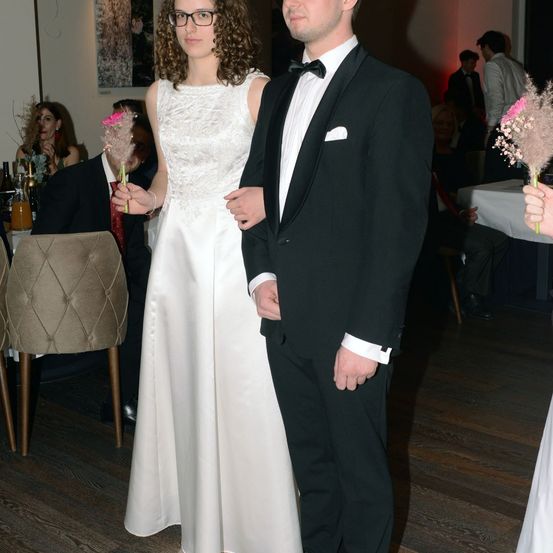 Ein Paar in formeller Kleidung bei einer Hochzeitsfeier, Blumen haltend, mit Gästen, die an Tischen im Hintergrund sitzen.