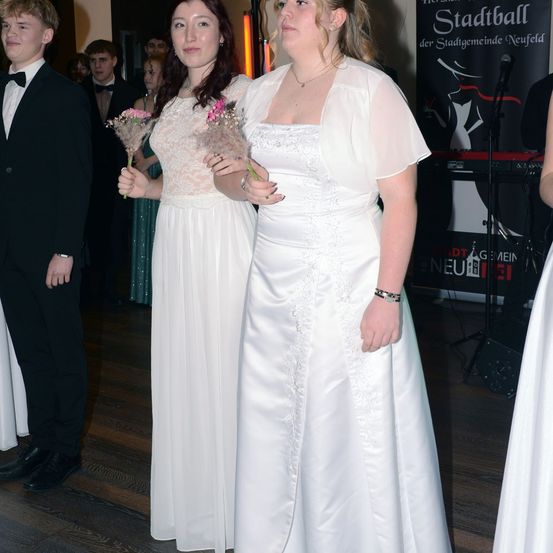 Zwei Frauen in weißen Kleidern halten Blumen in der Hand und stehen auf einem Holzboden, wahrscheinlich bei einer formellen Veranstaltung. Ein Mann in einem Anzug steht hinter ihnen, und ein Banner ist im Hintergrund zu sehen.