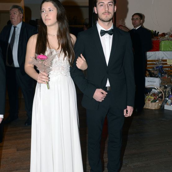 Ein Paar, eine junge Frau in einem weißen Kleid und ein junger Mann in einem schwarzen Frack, stehen zusammen in einem Raum. Dahinter sind verschwommene Figuren von Menschen zu sehen, einer trägt eine Brille.