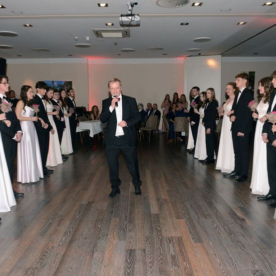 Ein Mann in einem schwarzen Anzug spricht in ein Mikrofon, während eine Gruppe von Menschen in formeller Kleidung um ihn herumsteht und Blumen hält. Die Veranstaltung scheint eine formelle Zusammenkunft oder Zeremonie in einem gut beleuchteten Raum mit Holzböden zu sein.