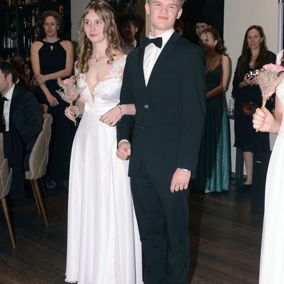 Ein junges Paar in formeller Kleidung steht in einem Raum mit Gästen. Die Frau hält rosa Blumen. Hinter ihnen sitzen und stehen Leute.