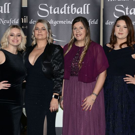 Vier Frauen in Abendkleidung stehen vor einem Banner mit 'Stadtball' darauf. Sie lächeln und scheinen für ein Foto zu posieren.