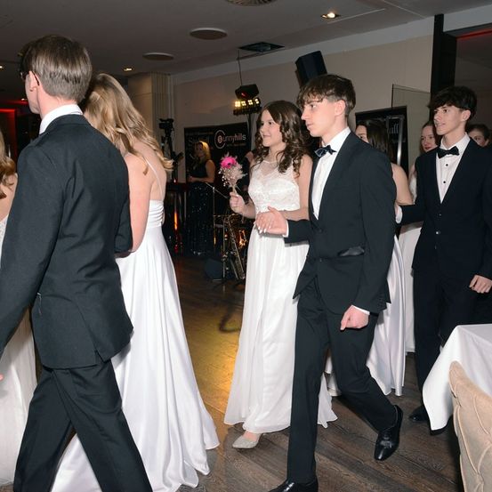 Eine Gruppe junger Menschen in formeller Kleidung geht gemeinsam in einem Saal. Ein Mädchen hält einen rosa Blumenstrauß. Eine Person vorne trägt eine Fliege.