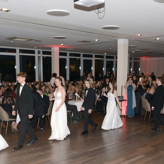 Eine Hochzeitsfeier mit Gästen, die an Tischen sitzen, zwei Brautjungfern und Trauzeugen, die auf der Tanzfläche gehen. Der Veranstaltungsort hat große Fenster und eine Decke mit Beleuchtung.