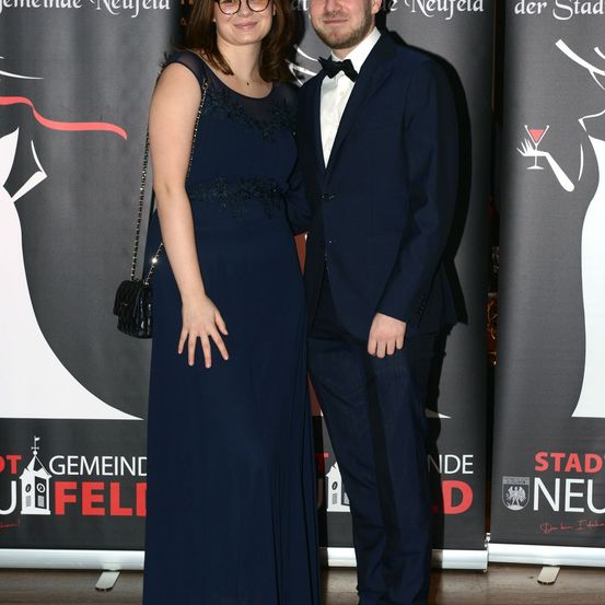 Ein Paar steht auf einem roten Teppich und lächelt. Die Frau trägt ein marineblaues Kleid und der Mann einen marineblauen Anzug. Sie stehen vor Bannern mit weißen Kleidern darauf.