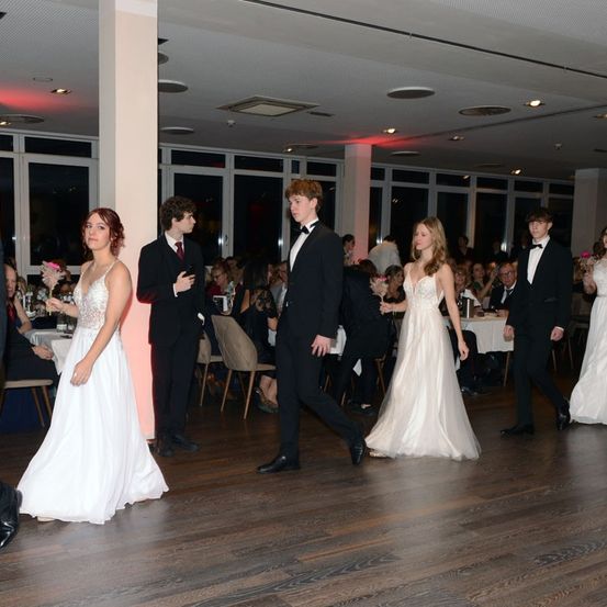 Eine Gruppe von Menschen in formeller Kleidung tanzt auf einem Holzboden in einem Saal. Einige Leute sitzen auf Stühlen um Tische. Es gibt viele Lampen an der Decke.