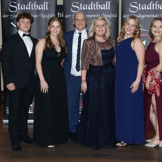 Fünf Personen posieren für ein Foto. Der Mann in der Mitte trägt eine Krawatte. Die Frauen tragen Kleider und hohe Schuhe. Die Frau auf der rechten Seite hält eine Handtasche. Hinter ihnen ist ein Banner mit dem Text 'Stadtball'.
