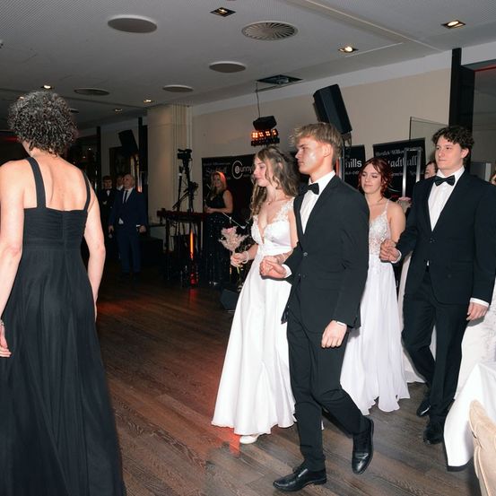 Eine Gruppe von Menschen in formeller Kleidung geht in einem Ballsaal. Eine Frau in einem schwarzen Kleid schaut zurück, während ein Mann in einem Anzug eine Frau in einem weißen Kleid führt.